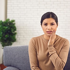 Woman in brown sweater sitting on couch and rubbing jaw